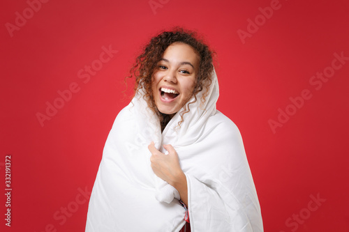 Cheerful young african american girl in pajamas homewear posing while resting at home isolated on red background in studio. Relax good mood lifestyle concept. Mock up copy space. Wrapping in blanket.