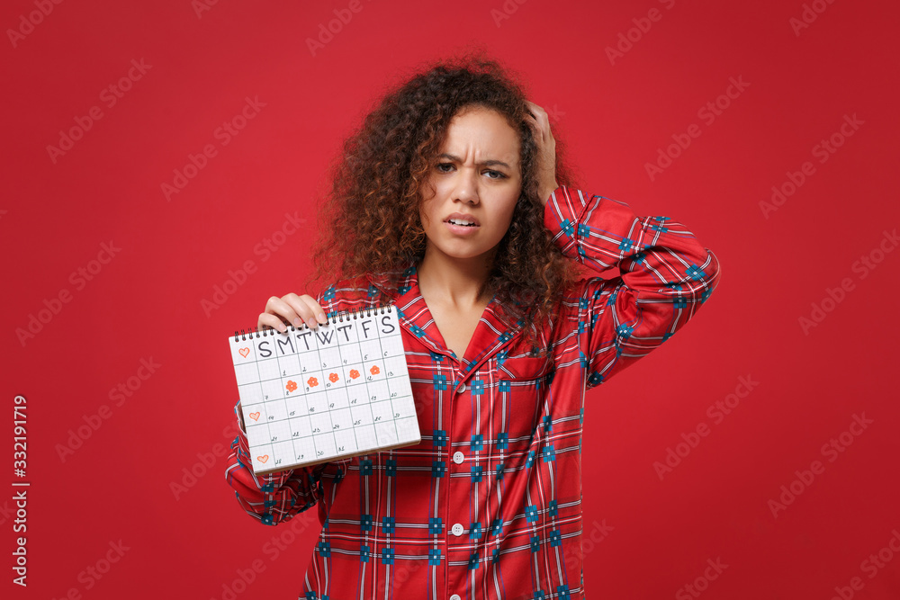Tired african american girl in pajamas homewear rest at home isolated on  red background. Relax good mood lifestyle concept Hold female periods  calendar for checking menstruation days put hand on head. Stock