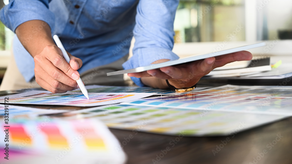 Cropped image of creative man writing/drawing/selecting a color from information color guide while working with computer tablet and color charts at the wooden working table over office as background. Cropped image of creative man writing/drawing/selecting a color from information color guide while working with computer tablet and color charts at the wooden working table over office as background.