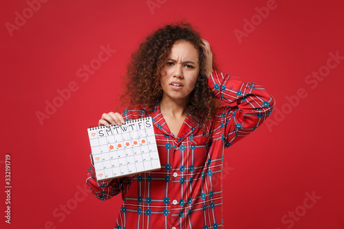 Tired african american girl in pajamas homewear rest at home isolated on red background. Relax good mood lifestyle concept Hold female periods calendar for checking menstruation days put hand on head. photo