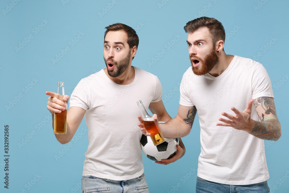 Shocked two young men guys friends in white t-shirt isolated on pastel blue background. Sport leisure concept. Cheer up support favorite team with soccer ball, beer bottle pointing index finger aside.