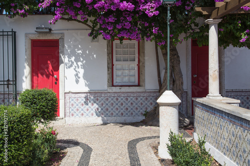 Fototapeta Naklejka Na Ścianę i Meble -  Lissabon, Portugal: Garten und Fassade mit typischen Fliesen, roter Tür  umrahmt von blühender lila Bougainvillea