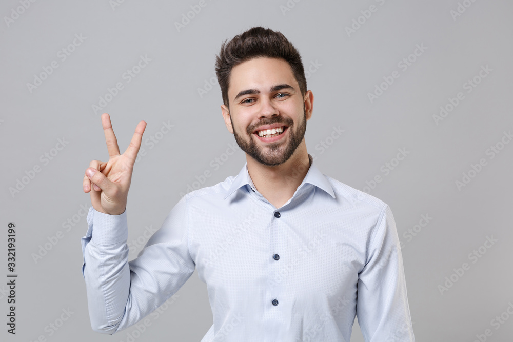 Cheerful young unshaven business man in light shirt posing isolated on grey wall background studio portrait. Achievement career wealth business concept. Mock up copy space. Showing victory sign.