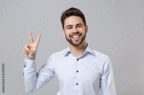 Cheerful young unshaven business man in light shirt posing isolated on grey wall background studio portrait. Achievement career wealth business concept. Mock up copy space. Showing victory sign.