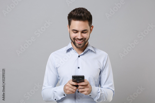 Smiling young unshaven business man in light shirt posing isolated on grey background in studio. Achievement career wealth business concept. Mock up copy space. Using mobile phone, typing sms message.