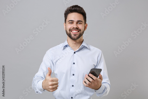 Smiling young unshaven business man in light shirt posing isolated on grey background in studio. Achievement career wealth business concept. Mock up copy space. Using mobile phone, showing thumb up.