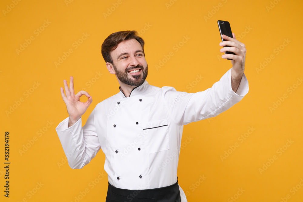 Smiling young bearded male chef cook or baker man in white uniform shirt isolated on yellow background. Cooking food concept. Mock up copy space. Doing selfie shot on mobile phone, showing OK gesture. Smiling young bearded male chef cook or baker man in white uniform shirt isolated on yellow background. Cooking food concept. Mock up copy space. Doing selfie shot on mobile phone, showing OK gesture.