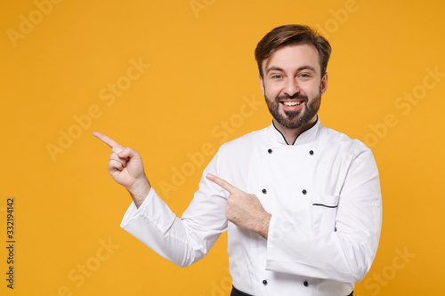 Funny young bearded male chef cook or baker man in white uniform shirt posing isolated on yellow wall background in studio. Cooking food concept. Mock up copy space. Pointing index fingers aside up.