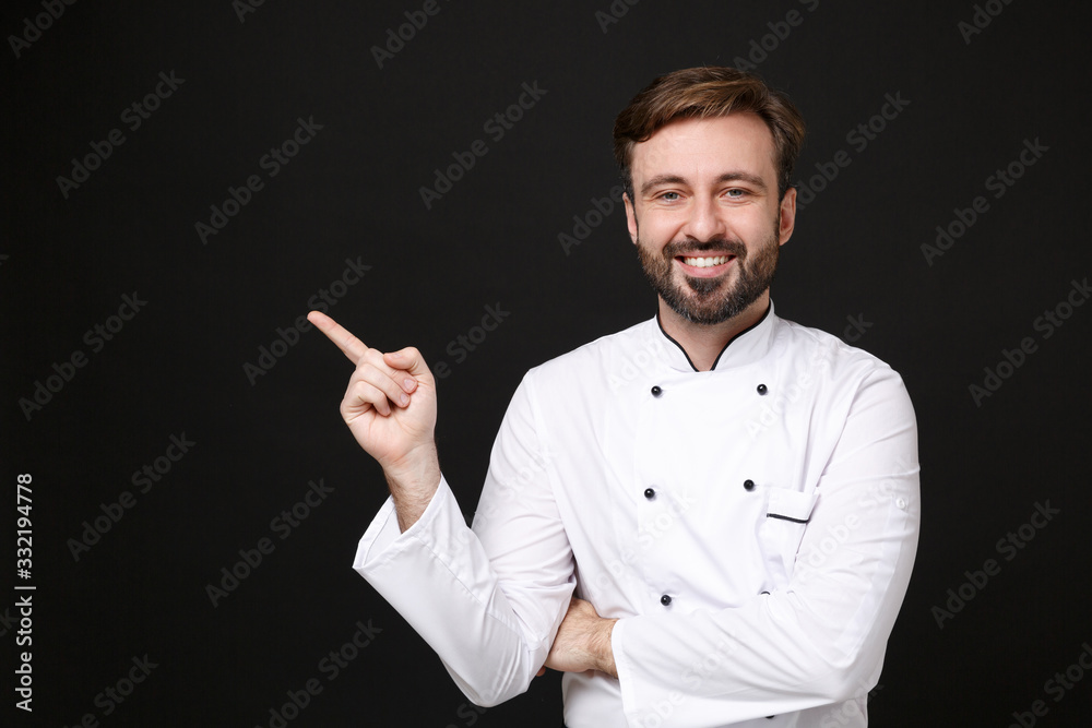 Smiling young bearded male chef cook or baker man in white uniform shirt posing isolated on black wall background studio portrait. Cooking food concept. Mock up copy space. Point index finger aside. Smiling young bearded male chef cook or baker man in white uniform shirt posing isolated on black wall background studio portrait. Cooking food concept. Mock up copy space. Point index finger aside.