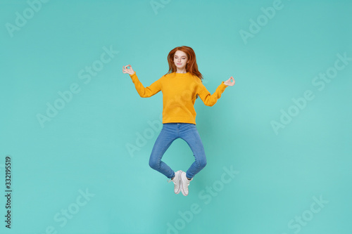 Young redhead woman girl in yellow sweater posing isolated on blue turquoise background in studio. People lifestyle concept. Mock up copy space. Jumping hold hands in yoga gesture relaxing meditating.