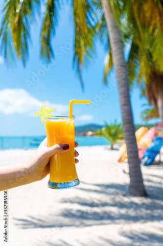 First-person view. Girl holds a glass cup of cold mango fresh on the background of a sandy tropical beach. White sand and palm trees. Fairytale vacation
