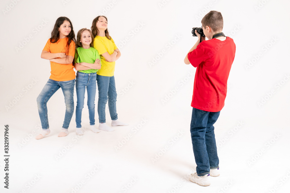 A boy photographer takes pictures of three cute girls in bright T-shirts on a white background in the studio. Young photographer and blogger. A boy photographer takes pictures of three cute girls in bright T-shirts on a white background in the studio. Young photographer and blogger.