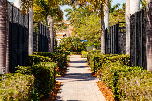 A paver brick pathway between a series of tennis courts leads to a clubhouse entrance. There are bushes between the tennis courts and the walkway giving a lush and tropical appearance.