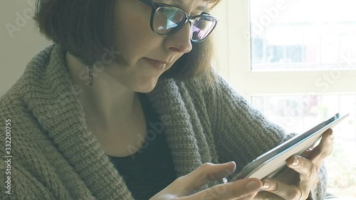 Young caucasian woman in cardigan and casual clothes wearing black frame eyeglasses and using tablet at home