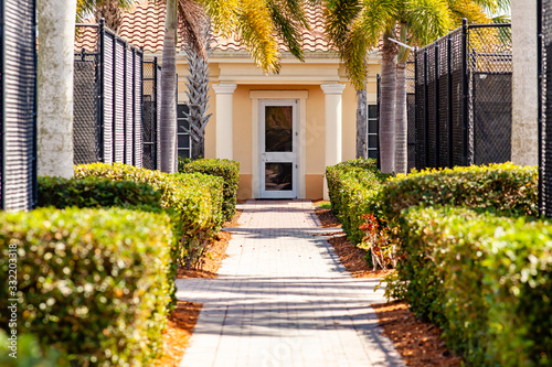 A brick pathway between a series of tennis courts leads to a clubhouse entrance. There are bushes between the tennis courts and the walkway giving a lush, tropical appearance