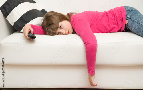 bored little girl lying on the sofa watches TV during the Coronavirus quarantine photo
