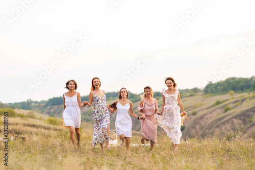 The company of cheerful female friends have a great time together on a picnic in a picturesque place overlooking the green hills. Girls in white dresses dancing in the field © Kate