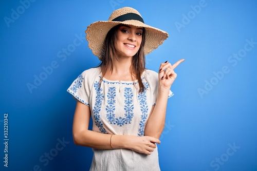 Young beautiful brunette woman wearing casual t-shirt and summer hat over blue background with a big smile on face, pointing with hand and finger to the side looking at the camera.