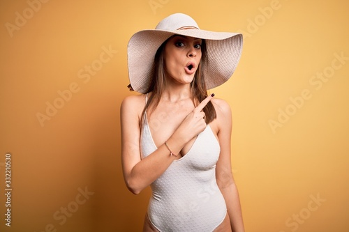 Young beautiful brunette woman on vacation wearing swimsuit and summer hat Surprised pointing with finger to the side, open mouth amazed expression.