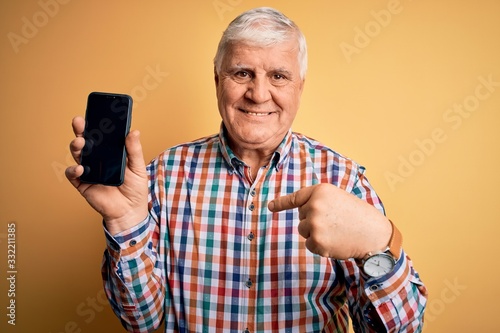 Senior handsome hoary man holding smartphone showing screen over yellow background with surprise face pointing finger to himself © Krakenimages.com