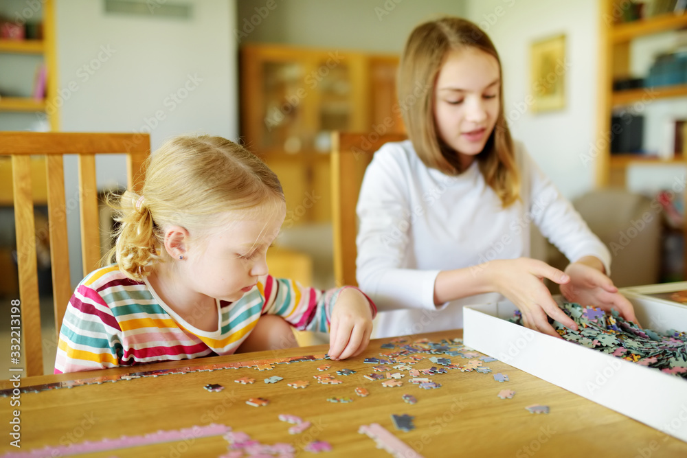 Fototapeta premium Cute young girls playing puzzles at home. Children connecting jigsaw puzzle pieces in a living room table. Kids assembling a jigsaw puzzle. Fun family leisure.