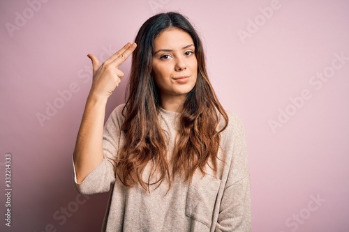 Young beautiful brunette woman wearing casual sweater standing over pink background Shooting and killing oneself pointing hand and fingers to head like gun, suicide gesture.