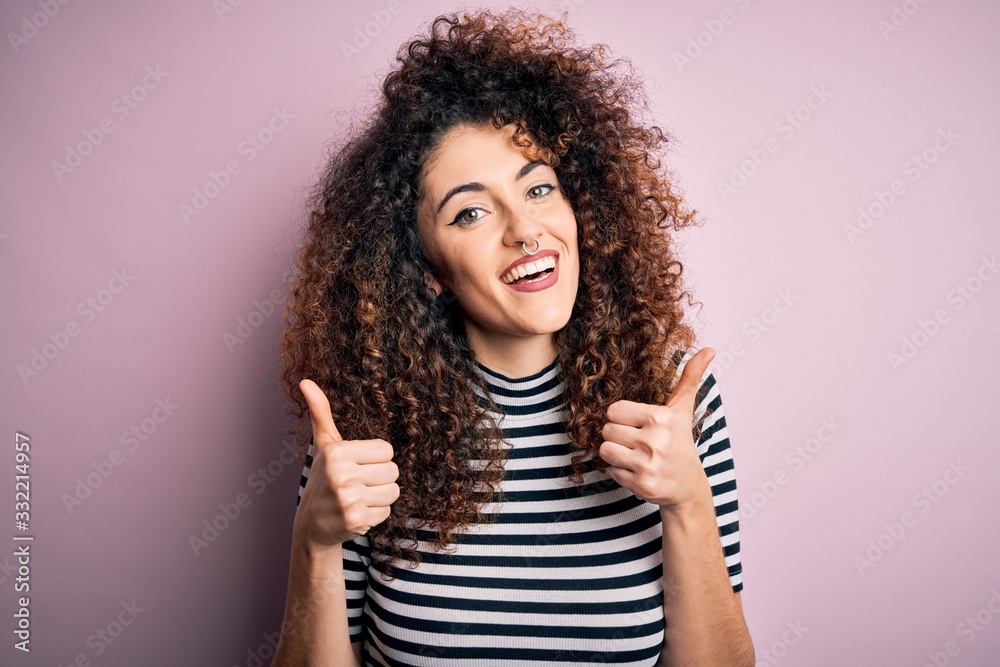 Young beautiful woman with curly hair and piercing wearing casual striped t-shirt success sign doing positive gesture with hand, thumbs up smiling and happy. Cheerful expression and winner gesture. Young beautiful woman with curly hair and piercing wearing casual striped t-shirt success sign doing positive gesture with hand, thumbs up smiling and happy. Cheerful expression and winner gesture.