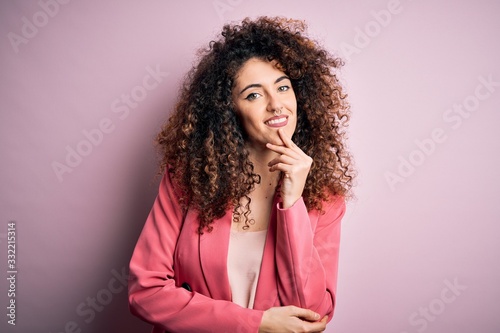 Young beautiful businesswoman with curly hair and piercing wearing elegant jacket looking confident at the camera smiling with crossed arms and hand raised on chin. Thinking positive. © Krakenimages.com