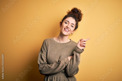 Young beautiful brunette woman with curly hair and piercing wearing casual dress with a big smile on face, pointing with hand and finger to the side looking at the camera.