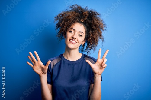 Young beautiful woman with curly hair and piercing wearing casual blue t-shirt showing and pointing up with fingers number eight while smiling confident and happy.
