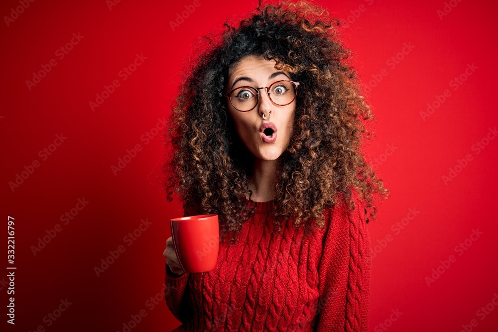 Young beautiful woman with curly hair and piercing drinking red cup of coffee scared in shock with a surprise face, afraid and excited with fear expression
