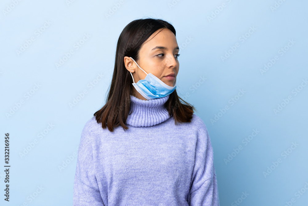 Young brunette mixed race woman protecting from the coronavirus with a mask and gloves over isolated blue background looking to the side Young brunette mixed race woman protecting from the coronavirus with a mask and gloves over isolated blue background looking to the side