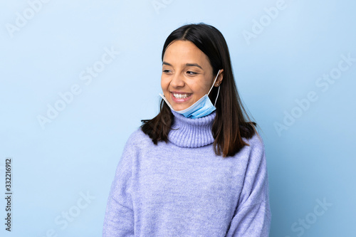 Young brunette mixed race woman protecting from the coronavirus with a mask and gloves over isolated blue background looking to the side and smiling
