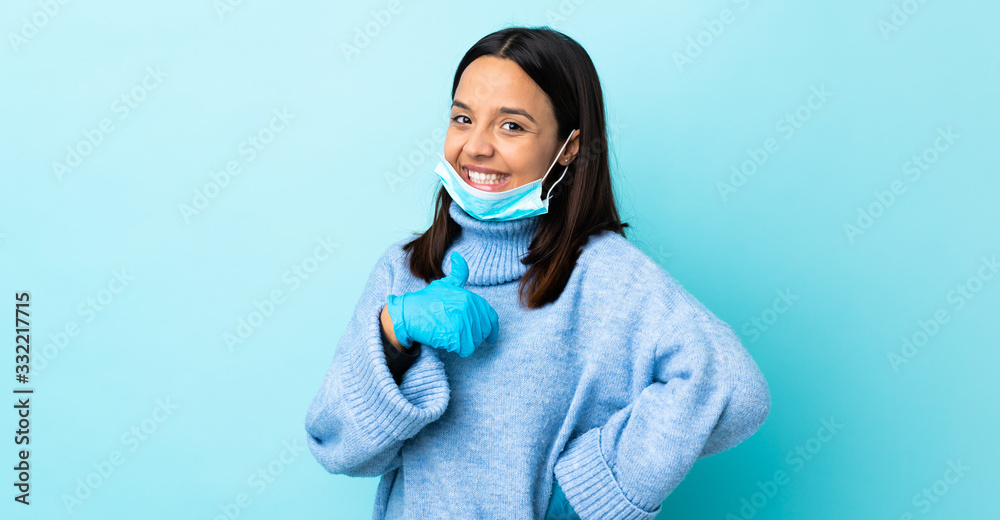 Young brunette mixed race woman protecting from the coronavirus with a mask and gloves over isolated blue background giving a thumbs up gesture Young brunette mixed race woman protecting from the coronavirus with a mask and gloves over isolated blue background giving a thumbs up gesture