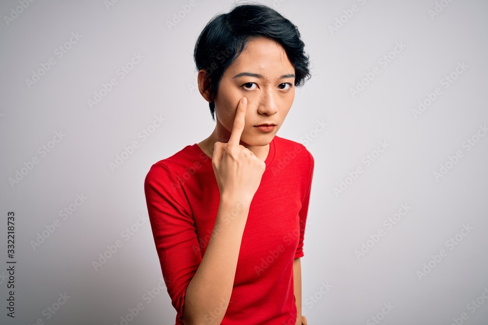 Young beautiful asian girl wearing casual red t-shirt standing over isolated white background Pointing to the eye watching you gesture, suspicious expression Young beautiful asian girl wearing casual red t-shirt standing over isolated white background Pointing to the eye watching you gesture, suspicious expression