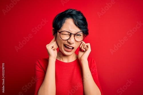 Young beautiful asian girl wearing casual t-shirt and glasses over isolated red background covering ears with fingers with annoyed expression for the noise of loud music. Deaf concept.