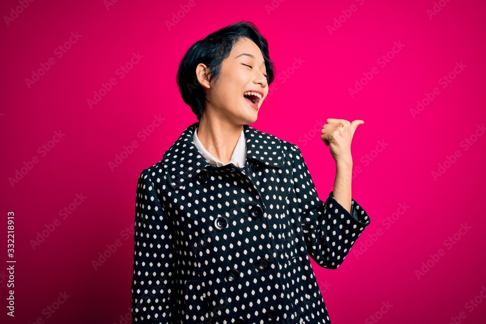 Young beautiful asian girl wearing casual jacket standing over isolated pink background smiling with happy face looking and pointing to the side with thumb up. Young beautiful asian girl wearing casual jacket standing over isolated pink background smiling with happy face looking and pointing to the side with thumb up.
