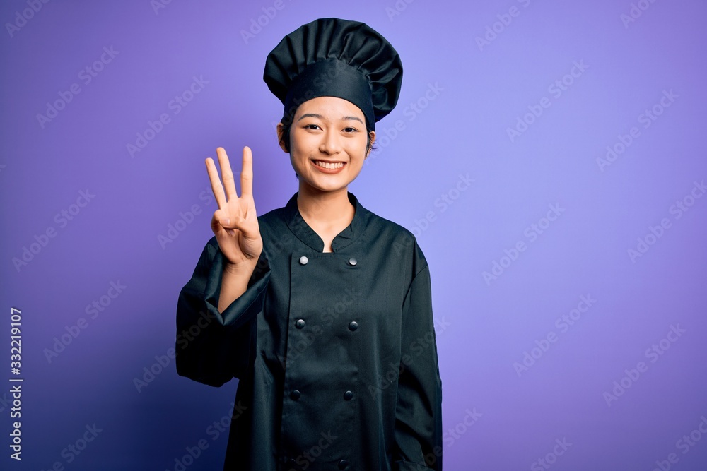 Young beautiful chinese chef woman wearing cooker uniform and hat over purple background showing and pointing up with fingers number three while smiling confident and happy. Young beautiful chinese chef woman wearing cooker uniform and hat over purple background showing and pointing up with fingers number three while smiling confident and happy.