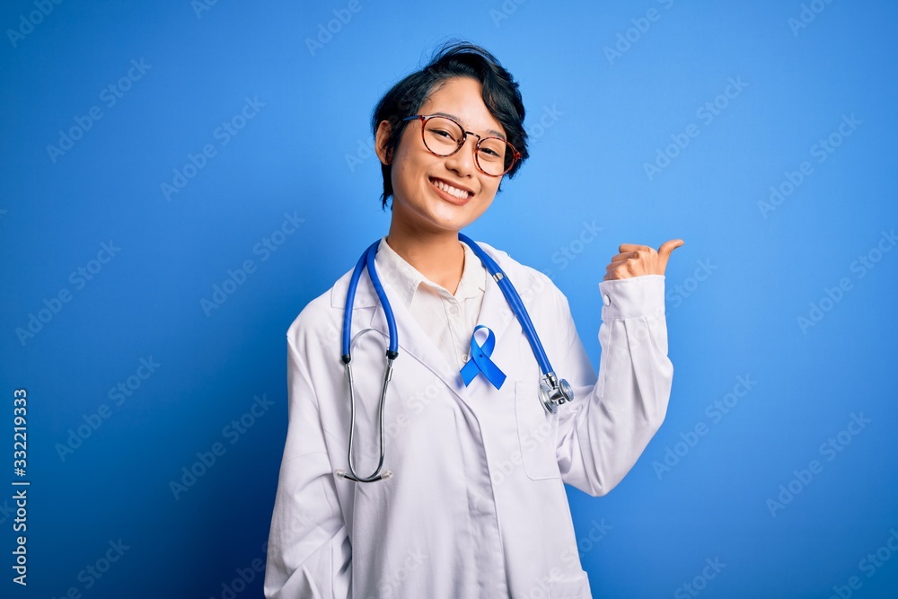 Young beautiful asian doctor girl wearing stethoscope and coat with blue cancer ribbon smiling with happy face looking and pointing to the side with thumb up. Young beautiful asian doctor girl wearing stethoscope and coat with blue cancer ribbon smiling with happy face looking and pointing to the side with thumb up.