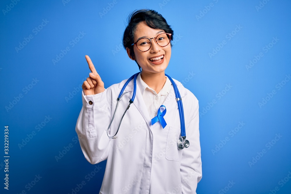 Young beautiful asian doctor girl wearing stethoscope and coat with blue cancer ribbon showing and pointing up with finger number one while smiling confident and happy. Young beautiful asian doctor girl wearing stethoscope and coat with blue cancer ribbon showing and pointing up with finger number one while smiling confident and happy.