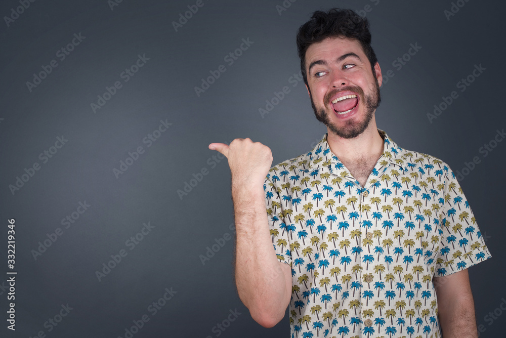 Omg concept. Stupefied caucasian male with surprised expression, opens eyes and mouth widely, points aside with thumb, shows something strange on gray background. Advertisement concept.