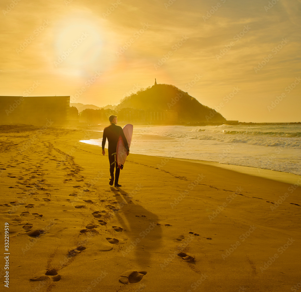 Fototapeta premium A surfer walking on the Zurriola Beach at sunset with the Monte Urgull in the background. San Sebastian, Basque Country, Guipuzcoa. Spain.