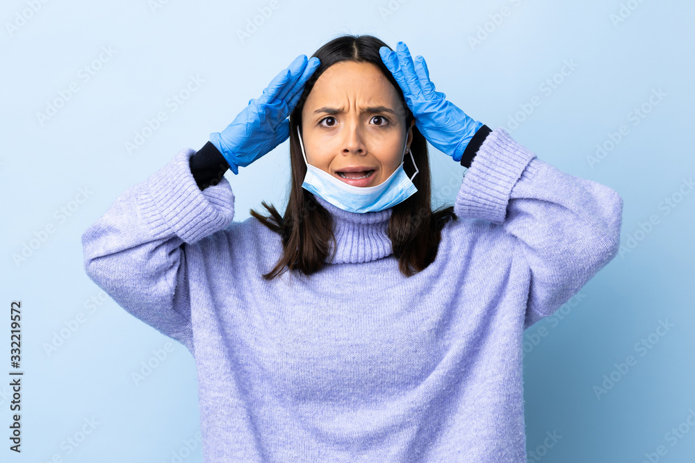 Young brunette mixed race woman protecting from the coronavirus with a mask and gloves over isolated blue background with surprise expression Young brunette mixed race woman protecting from the coronavirus with a mask and gloves over isolated blue background with surprise expression