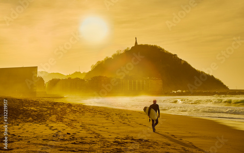 A surfer walking on the Zurriola Beach at sunset with the Monte Urgull in the background. San Sebastian, Basque Country, Guipuzcoa. Spain. photo