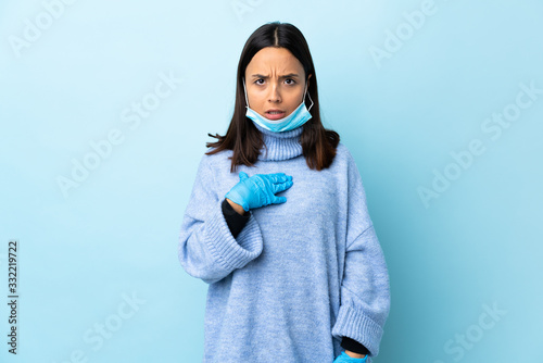 Young brunette mixed race woman protecting from the coronavirus with a mask and gloves over isolated blue background pointing to oneself