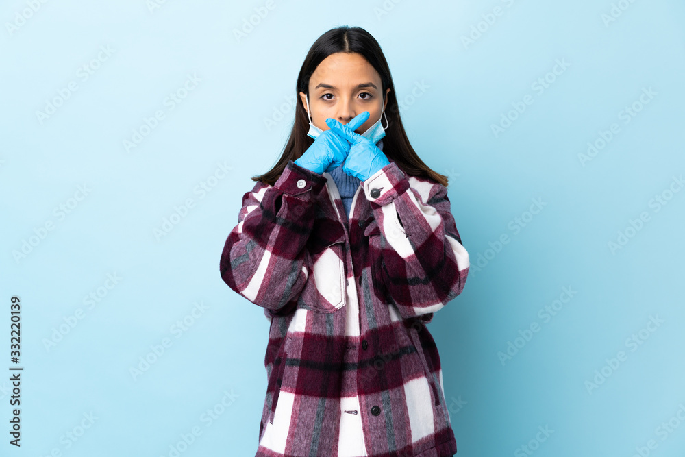 Young brunette mixed race woman protecting from the coronavirus with a mask and gloves over isolated blue background showing a sign of silence gesture Young brunette mixed race woman protecting from the coronavirus with a mask and gloves over isolated blue background showing a sign of silence gesture