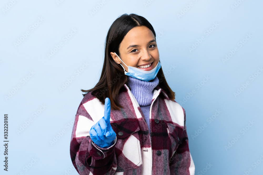 Young brunette mixed race woman protecting from the coronavirus with a mask and gloves over isolated blue background showing and lifting a finger Young brunette mixed race woman protecting from the coronavirus with a mask and gloves over isolated blue background showing and lifting a finger