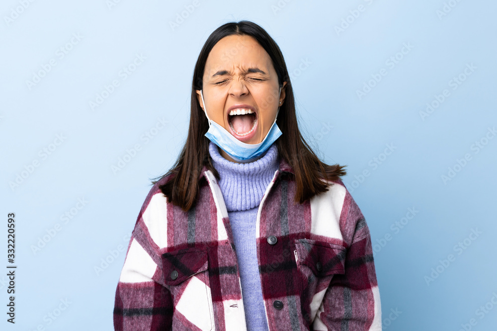 Young brunette mixed race woman protecting from the coronavirus with a mask and gloves over isolated blue background shouting to the front with mouth wide open