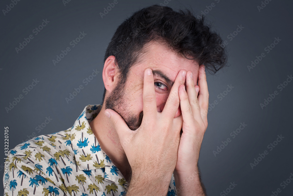 Anxiety - a conceptual image of a beautiful young caucasian man covering his face with his hands and peering out with one eye between her fingers standing indoors. Scared from something or someone. Anxiety - a conceptual image of a beautiful young caucasian man covering his face with his hands and peering out with one eye between her fingers standing indoors. Scared from something or someone.