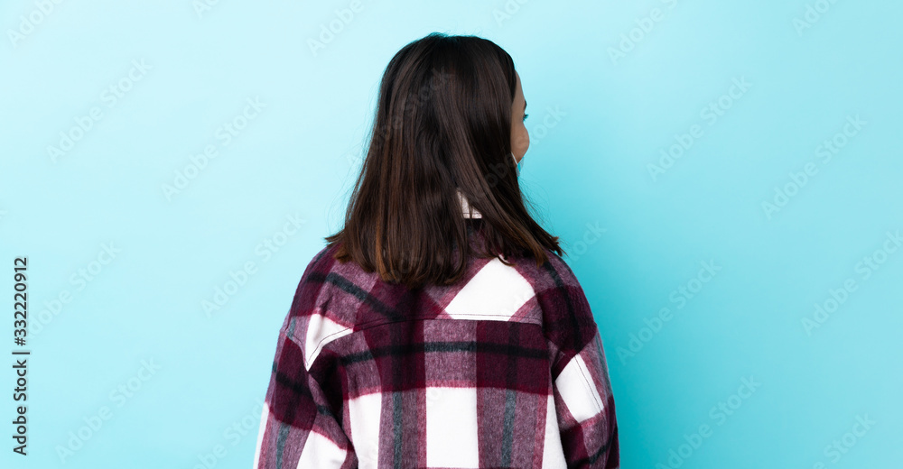 Young brunette mixed race woman protecting from the coronavirus with a mask and gloves over isolated blue background in back position and looking side Young brunette mixed race woman protecting from the coronavirus with a mask and gloves over isolated blue background in back position and looking side